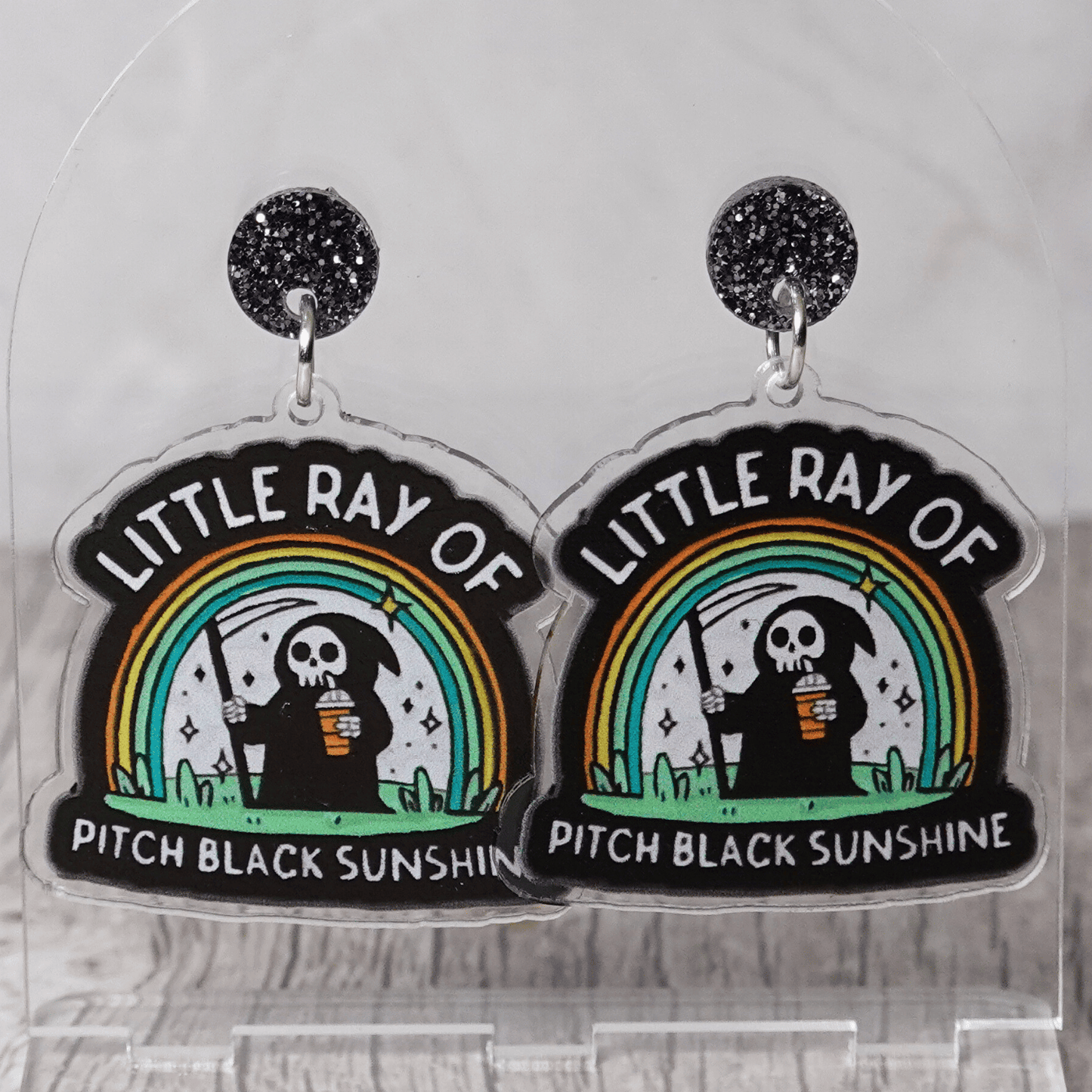 Novelty dangle earrings with a cartoon grim reaper holding a drink under a rainbow and the text “Little ray of pitch black sunshine”