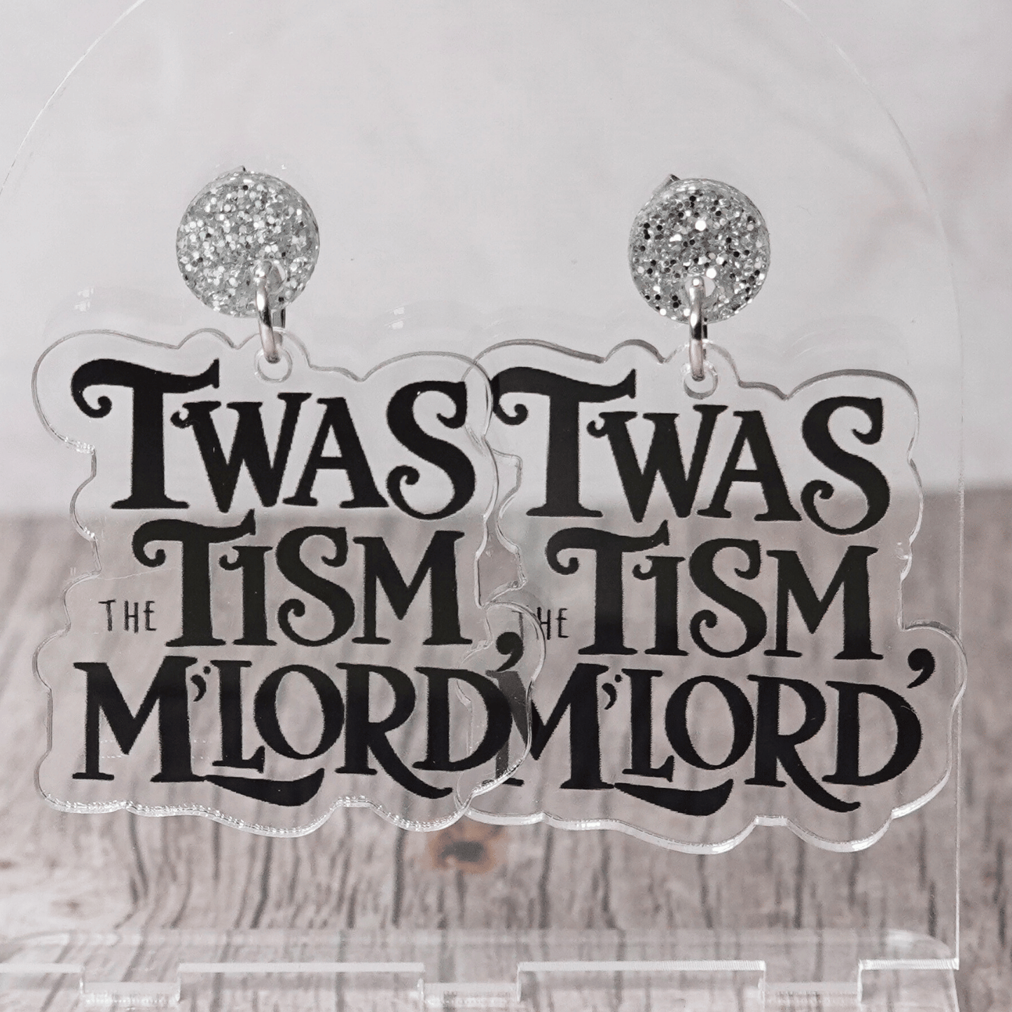 Clear acrylic dangle earrings with black text reading “‘Twas the tism, m’lord” in decorative lettering