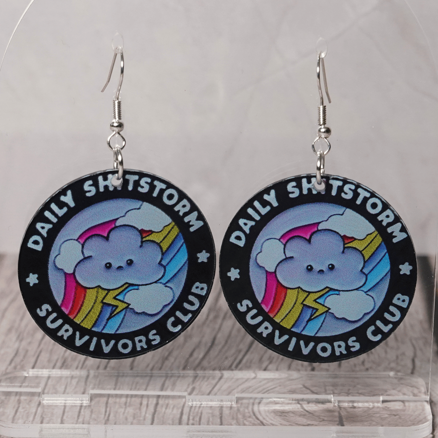 Round novelty dangle earrings reading “Daily Shitstorm Survivors Club” with a cute cloud, rainbow and lightning design
