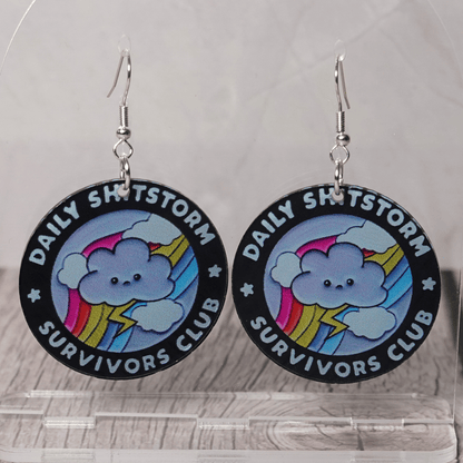 Round novelty dangle earrings reading “Daily Shitstorm Survivors Club” with a cute cloud, rainbow and lightning design