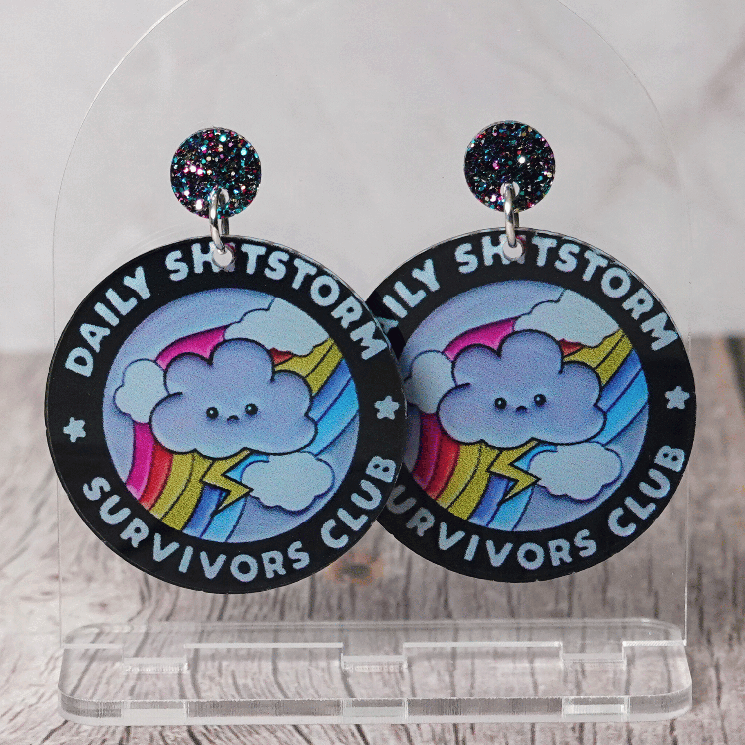 Round novelty dangle earrings reading “Daily Shitstorm Survivors Club” with a cute cloud, rainbow and lightning design