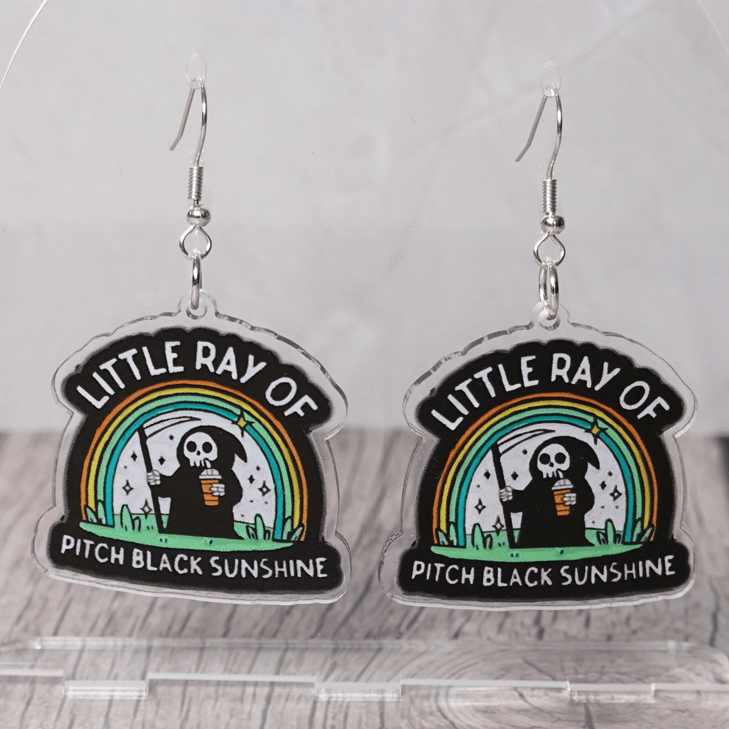 Novelty dangle earrings with a cartoon grim reaper holding a drink under a rainbow and the text “Little ray of pitch black sunshine”
