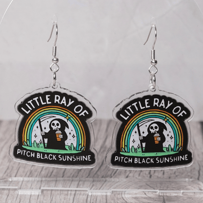 Novelty dangle earrings with a cartoon grim reaper holding a drink under a rainbow and the text “Little ray of pitch black sunshine”