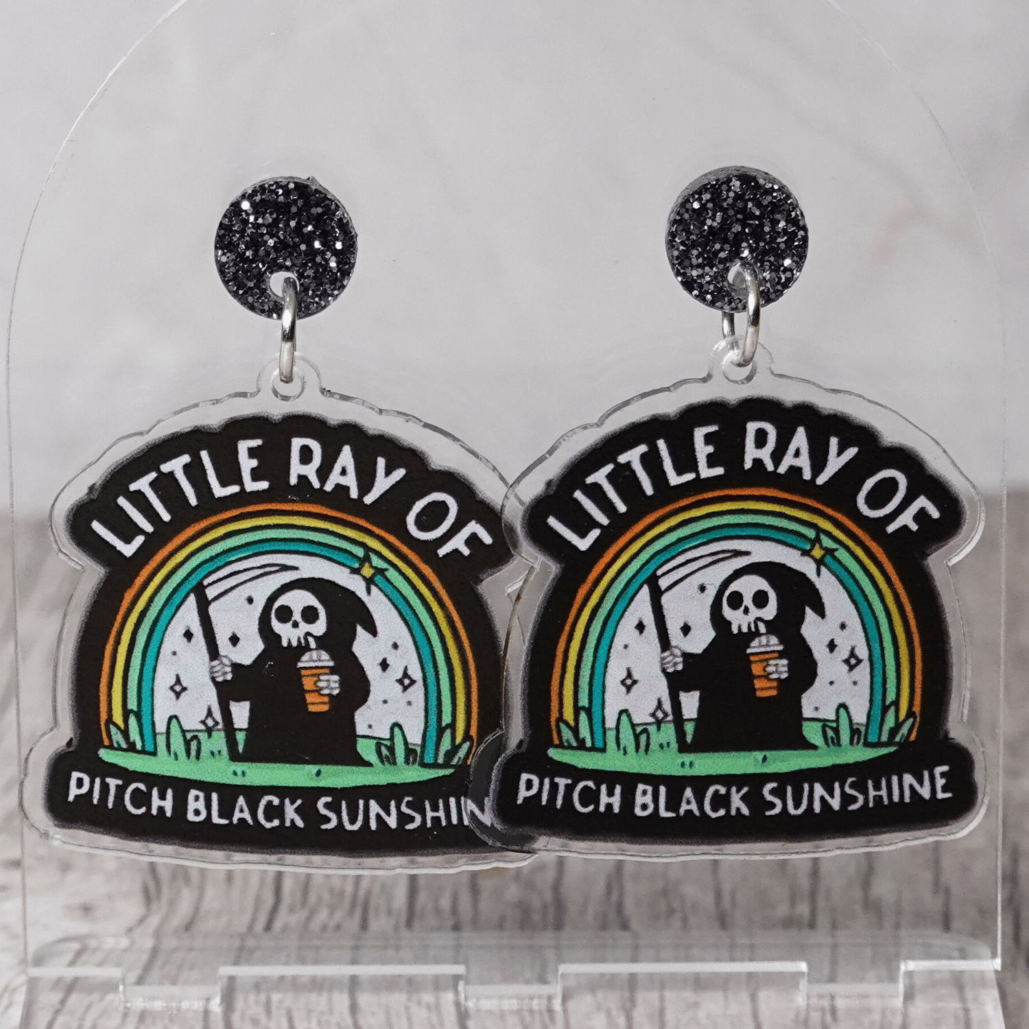 Novelty dangle earrings with a cartoon grim reaper holding a drink under a rainbow and the text “Little ray of pitch black sunshine”