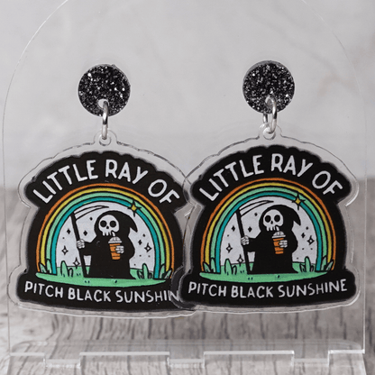Novelty dangle earrings with a cartoon grim reaper holding a drink under a rainbow and the text “Little ray of pitch black sunshine”