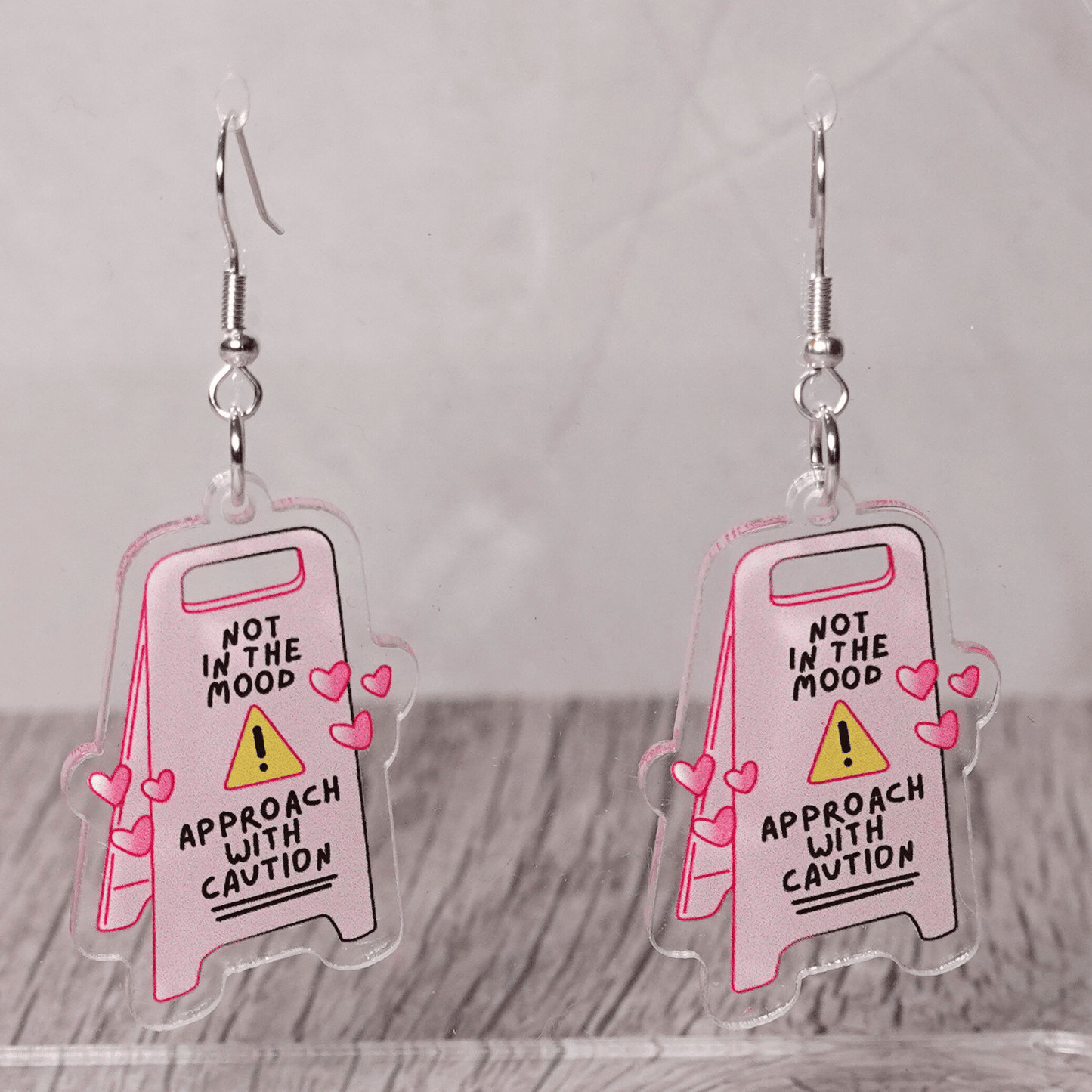 Funny acrylic dangle earrings shaped like a caution sign that reads “Not in the mood – approach with caution,” decorated with small pink hearts.