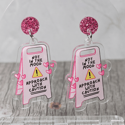Funny acrylic dangle earrings shaped like a caution sign that reads “Not in the mood – approach with caution,” decorated with small pink hearts.