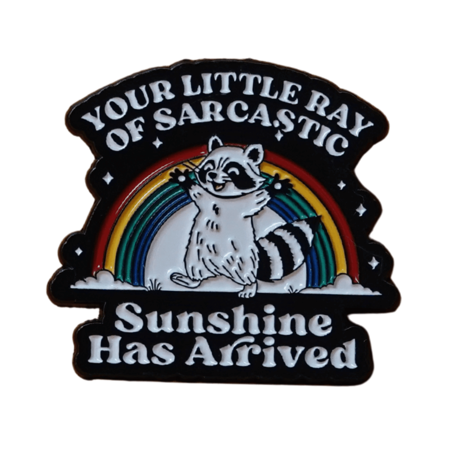 Enamel pin featuring a raccoon in front of a rainbow with the quote “Your little ray of sarcastic sunshine has arrived.”