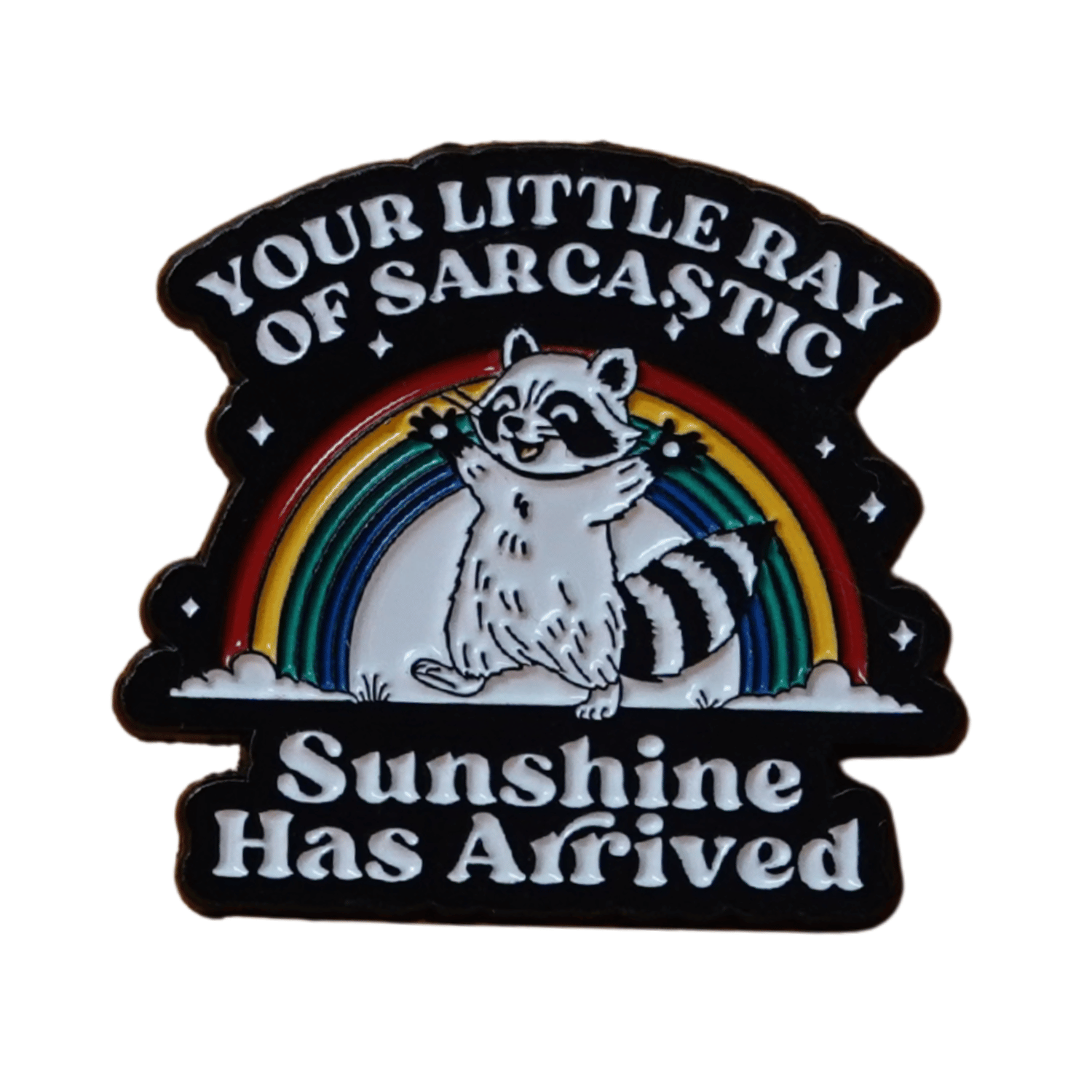 Enamel pin featuring a raccoon in front of a rainbow with the quote “Your little ray of sarcastic sunshine has arrived.”