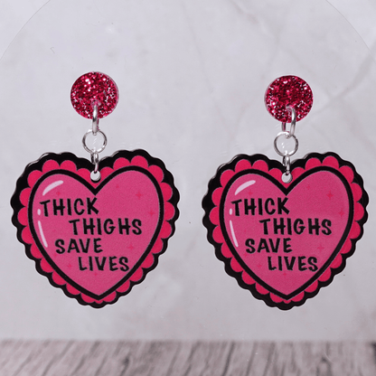 Pink heart dangle earrings with text “thick thighs save lives” and glitter stud tops
