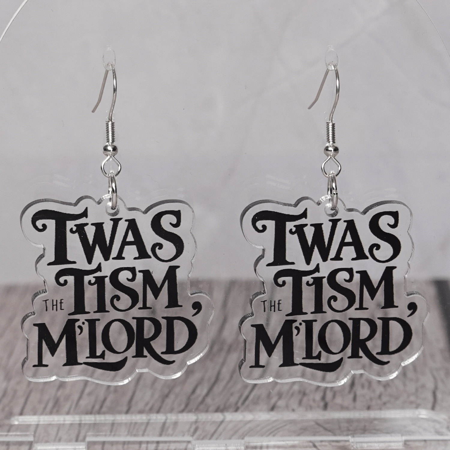 Clear acrylic dangle earrings with black text reading “‘Twas the tism, m’lord” in decorative lettering