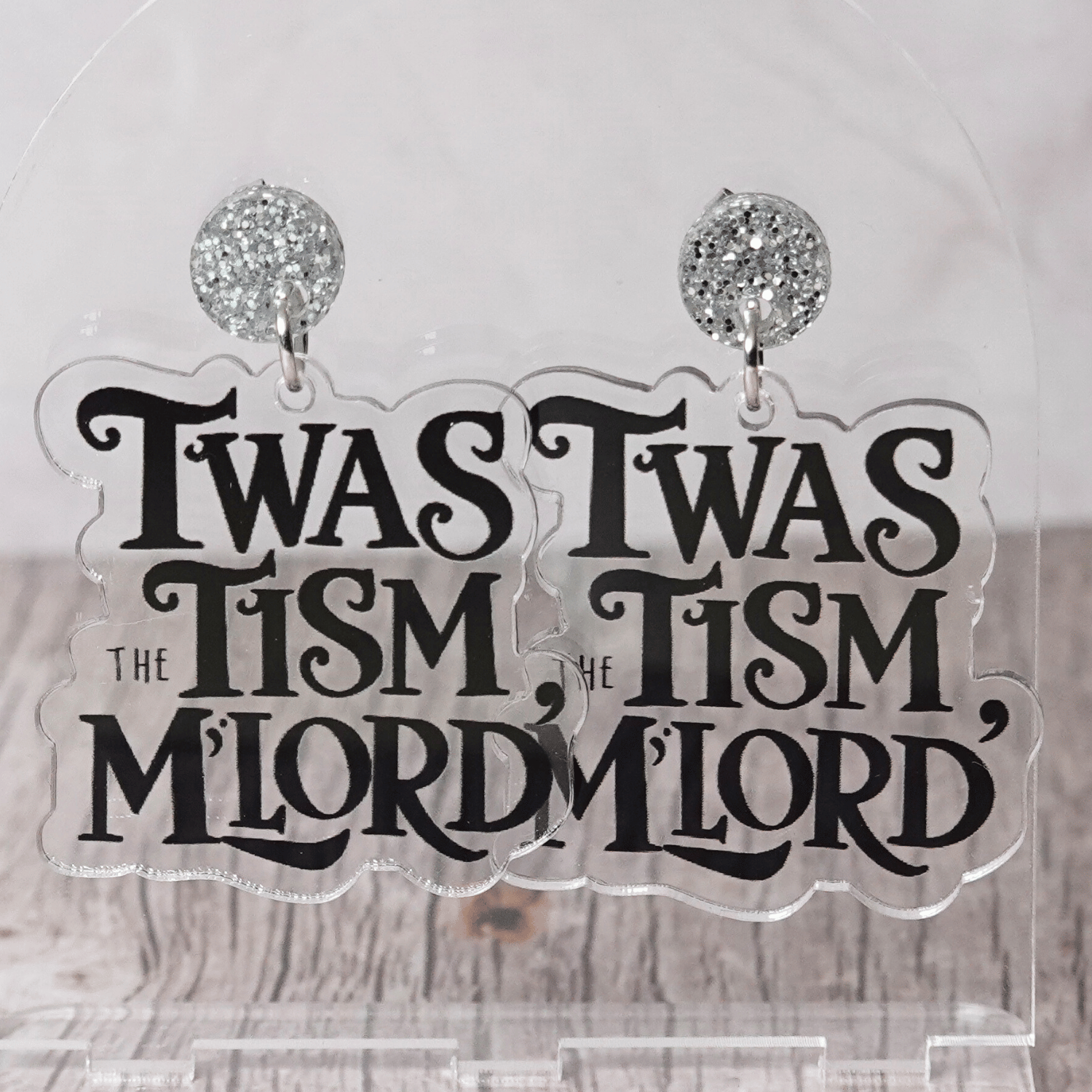 Clear acrylic dangle earrings with black text reading “‘Twas the tism, m’lord” in decorative lettering