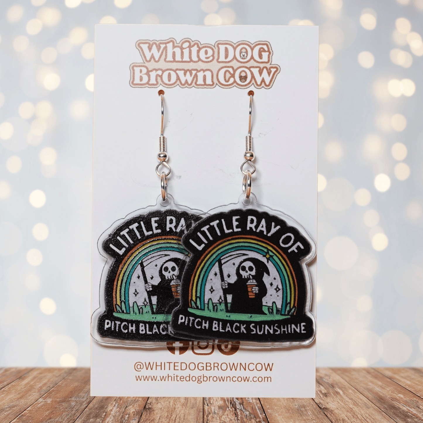 Novelty dangle earrings with a cartoon grim reaper holding a drink under a rainbow and the text “Little ray of pitch black sunshine”