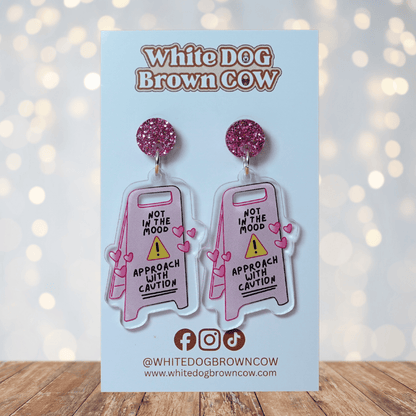 Funny acrylic dangle earrings shaped like a caution sign that reads “Not in the mood – approach with caution,” decorated with small pink hearts.