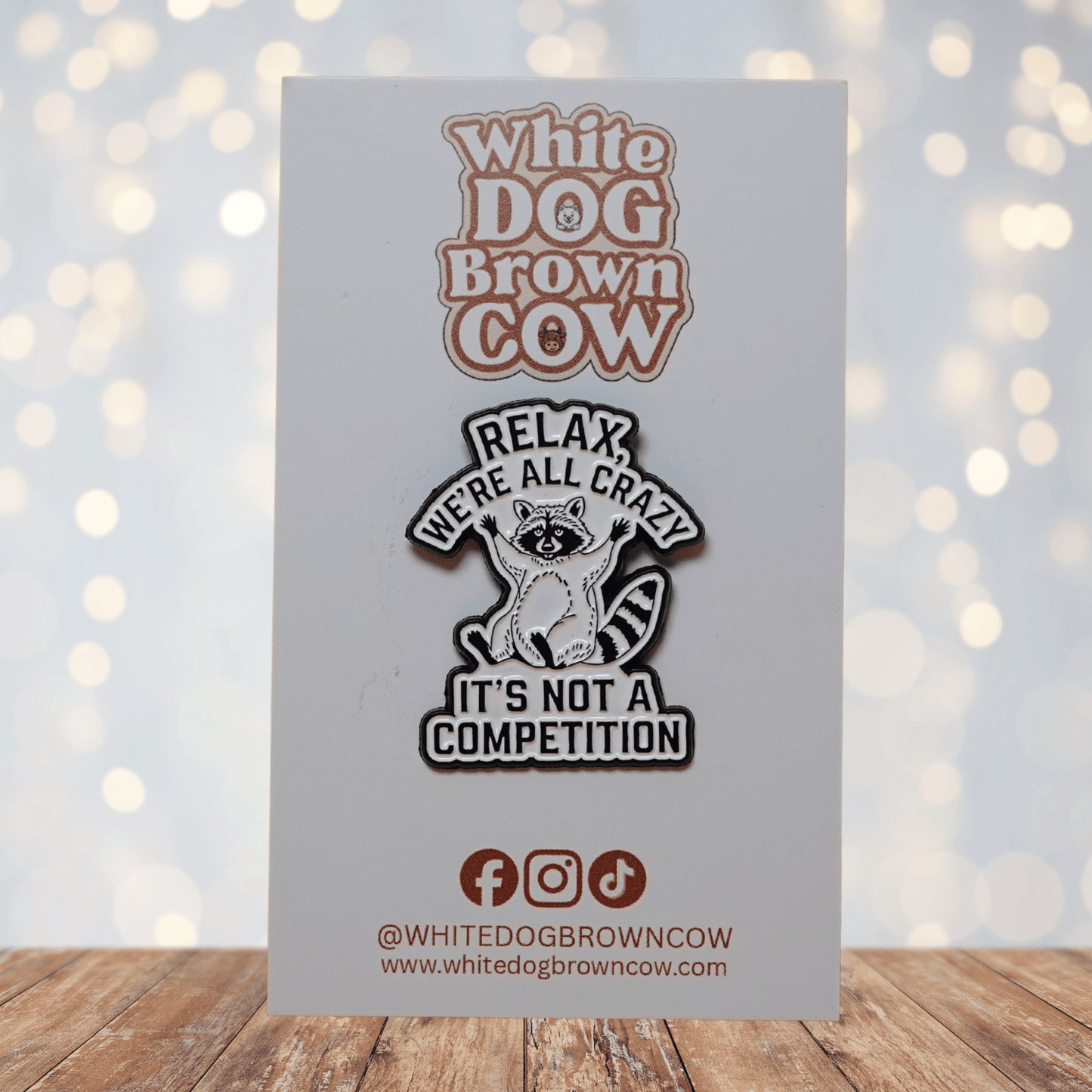 Black and white enamel pin with raccoon illustration and quote “Relax, We’re All Crazy, It’s Not a Competition,” funny sarcastic lapel pin.