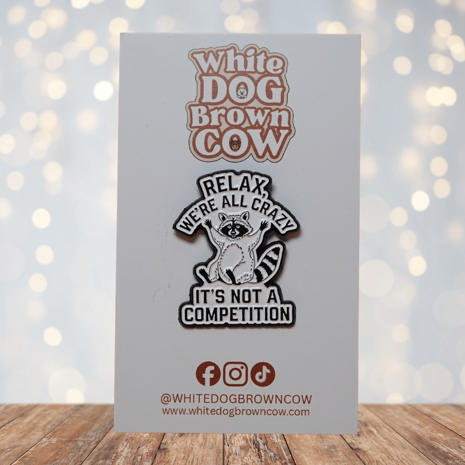 Black and white enamel pin with raccoon illustration and quote “Relax, We’re All Crazy, It’s Not a Competition,” funny sarcastic lapel pin.