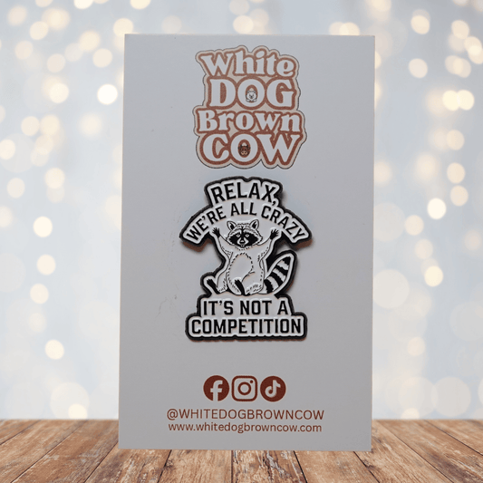 Black and white enamel pin with raccoon illustration and quote “Relax, We’re All Crazy, It’s Not a Competition,” funny sarcastic lapel pin.