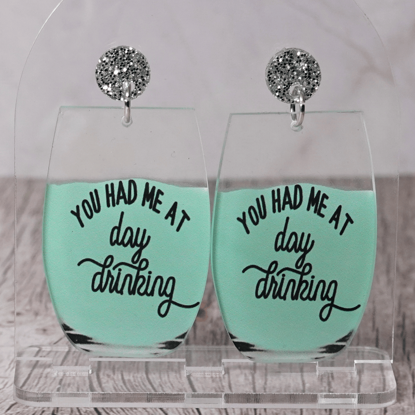 Funny acrylic dangle earrings shaped like wine glasses with mint green drink and text that reads “You had me at day drinking.”